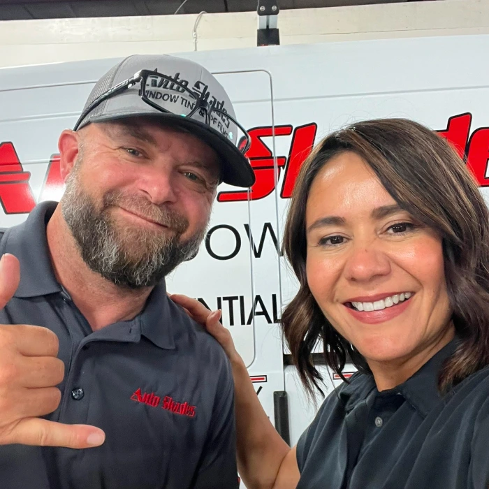 Two smiling Auto Shades team members pose for a selfie in front of a branded company van. The man wears a logo hat and makes a shaka hand gesture, while the woman stands beside him with her hand on his shoulder.
