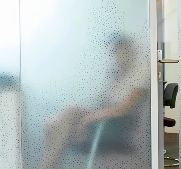 Frosted glass office partition with a subtle dotted floral pattern, partially obscuring the view of a seated person behind it, with a chair and door handle visible on the outside.