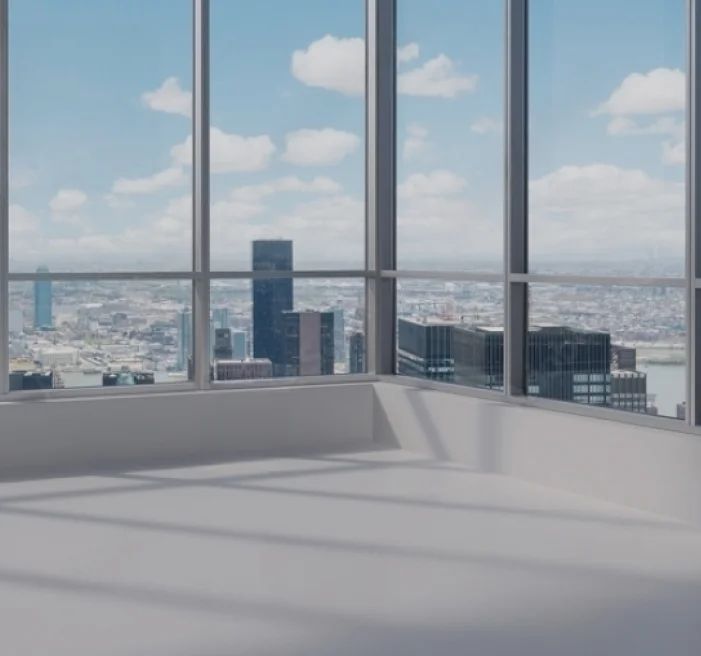 Corner of a high-rise room with floor-to-ceiling glass windows offering a panoramic view of a city skyline filled with skyscrapers and a river under a partly cloudy sky.