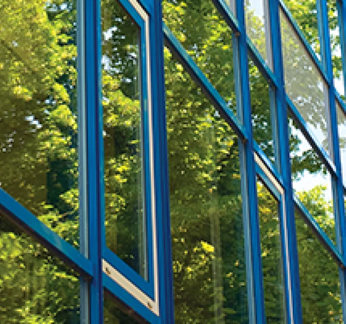 Close-up of a modern building facade with blue-framed glass windows reflecting green trees and sunlight. One window is slightly open, adding depth and dimension to the geometric structure.