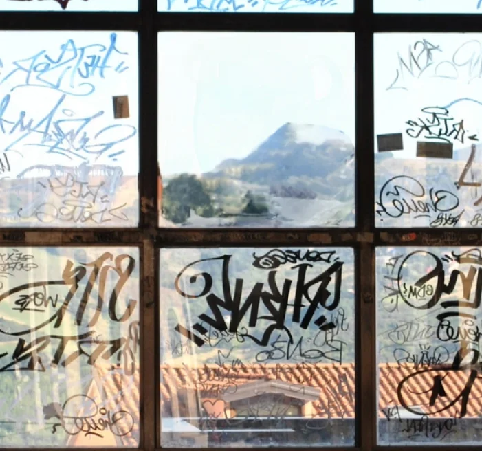 Multi-pane window covered in graffiti tags and markings, partially obscuring a scenic view of red tile rooftops, trees, and a distant mountain under a clear sky.