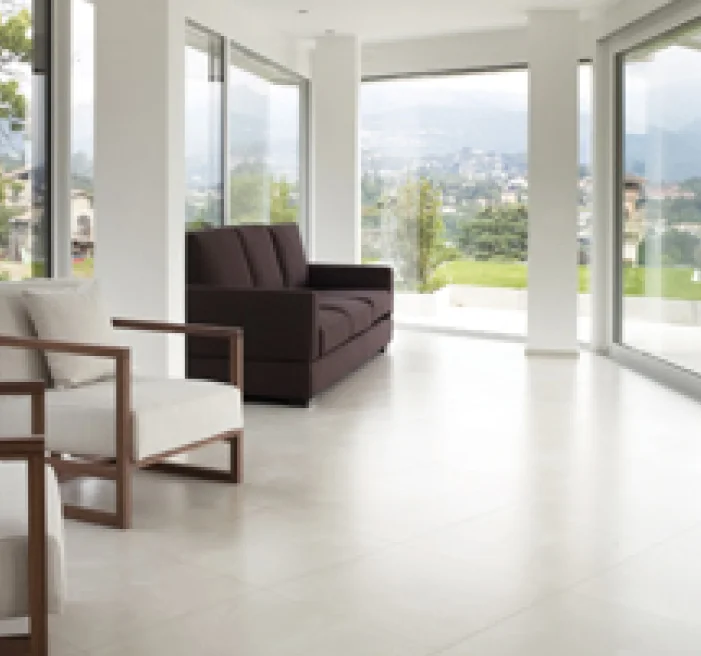 Bright and minimalistic living space with large glass walls offering sweeping views of a lush green landscape and distant mountains, furnished with a brown sofa and modern white chairs with wooden frames.