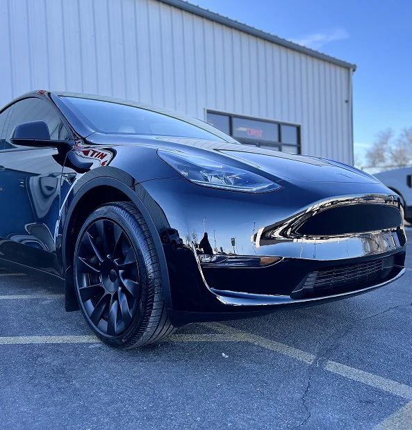 Shiny black Tesla Model Y with tinted windows and black wheels parked outside a commercial building. The car's glossy finish reflects nearby structures and a clear blue sky.