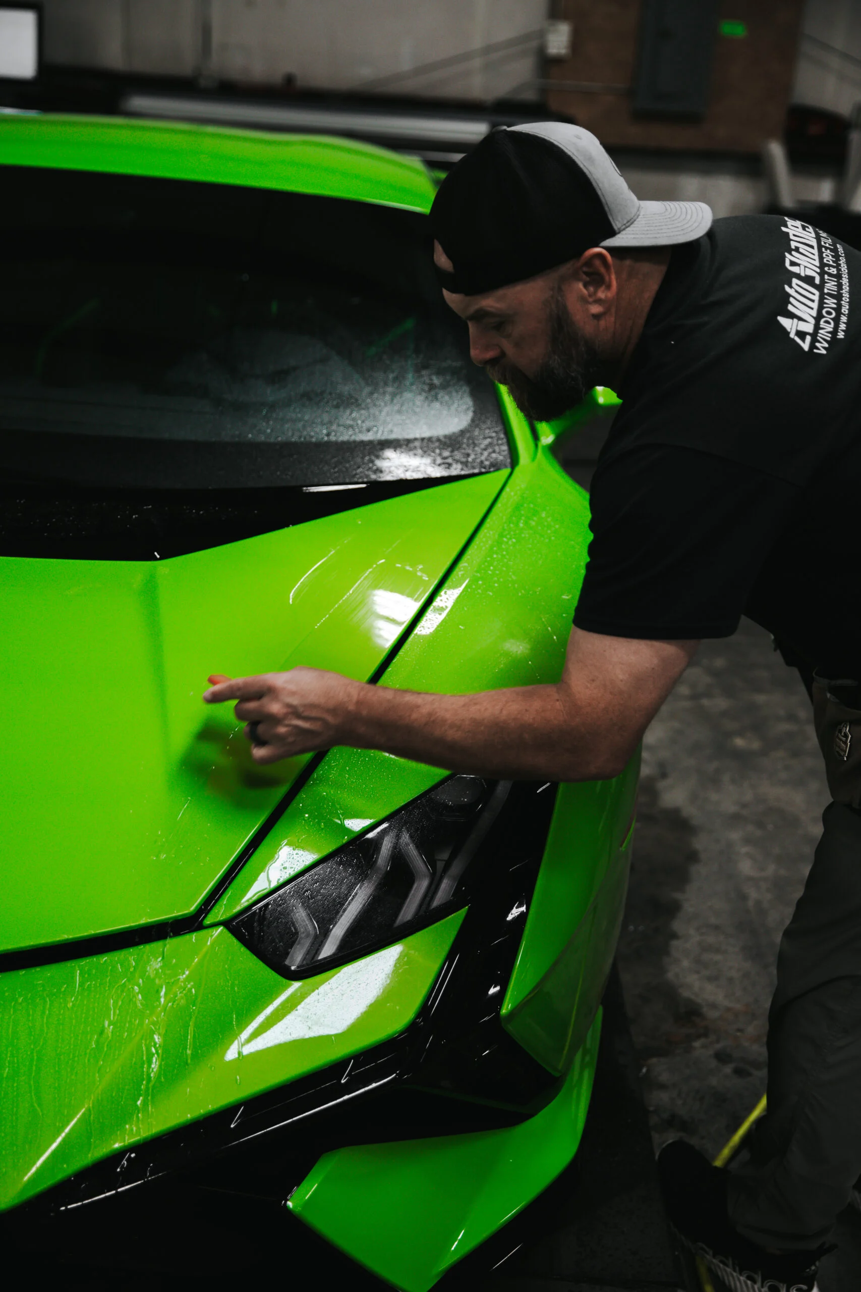A technician from Auto Shades carefully applies paint protection film to the hood of a bright green sports car, smoothing it out with a squeegee tool.