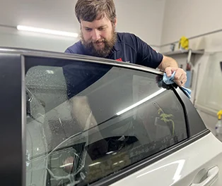 Technician carefully applies window tint film to the rear passenger window of a white vehicle inside a workshop.