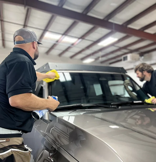 Two technicians clean and prep the windshield of a gray SUV inside a garage, using yellow microfiber cloths and spray cleaner.