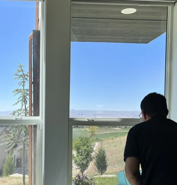 Man applying window film inside a home with large windows overlooking a scenic outdoor view of trees, grassy hills, and distant mountains under a clear blue sky.