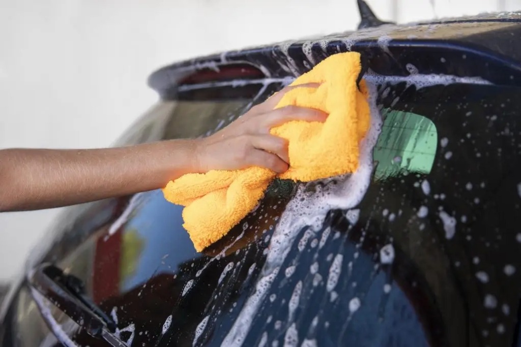A hand uses a bright yellow microfiber cloth to gently clean a tinted car window with soapy water, demonstrating proper maintenance techniques for preserving tint quality—an essential part of understanding What Window Tinting Really Is.