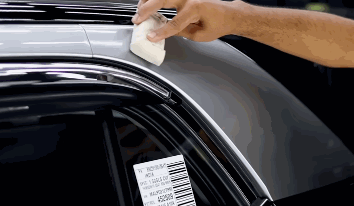 A person applies ceramic coating to a car's roof rail using a white applicator pad, near a barcode label on the vehicle’s door frame.