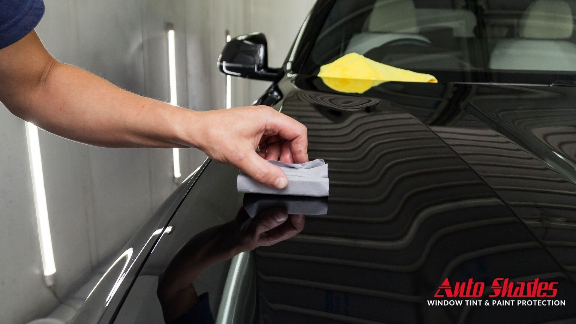 A hand using a folded microfiber cloth to apply ceramic coating on a glossy black car hood inside a detailing booth, with strong light reflections visible.