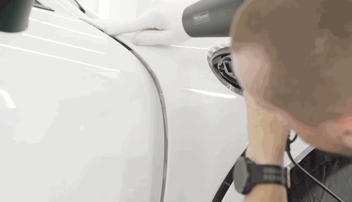 A close-up of a technician using a heat gun and gloved hands to seal paint protection film along the edge of a car's fuel cap area.