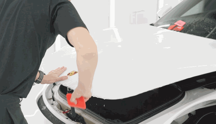 A technician installs paint protection film on the hood of a white Porsche, smoothing it with a red squeegee to remove water and air bubbles.