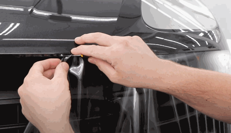 Close-up of hands using a tool to precisely apply paint protection film to the front edge of a car's hood and fender area.