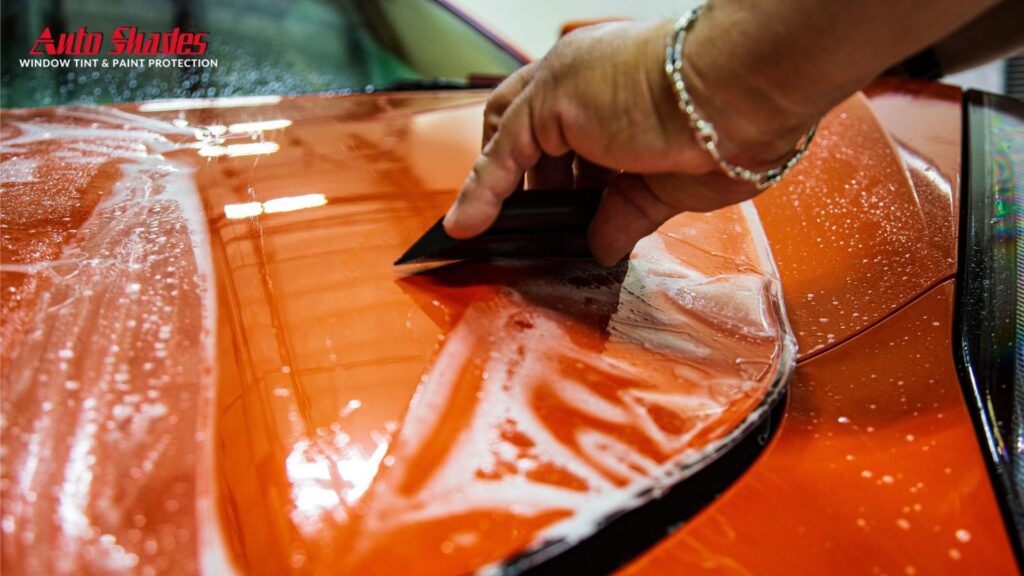 A person uses an applicator pad to smooth paint protection film onto the front fender of a silver car, with the Auto Shades logo in the corner.