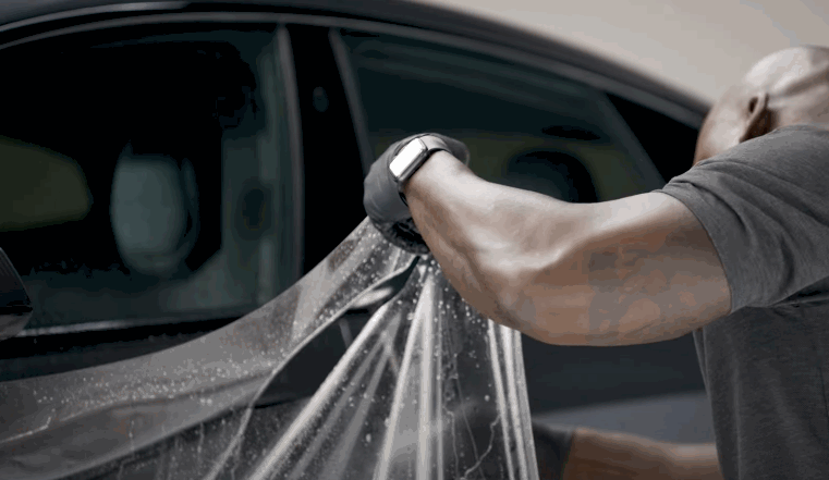 A person wearing gloves applies a wet sheet of paint protection film to a car door, carefully smoothing it out against the glass and frame.