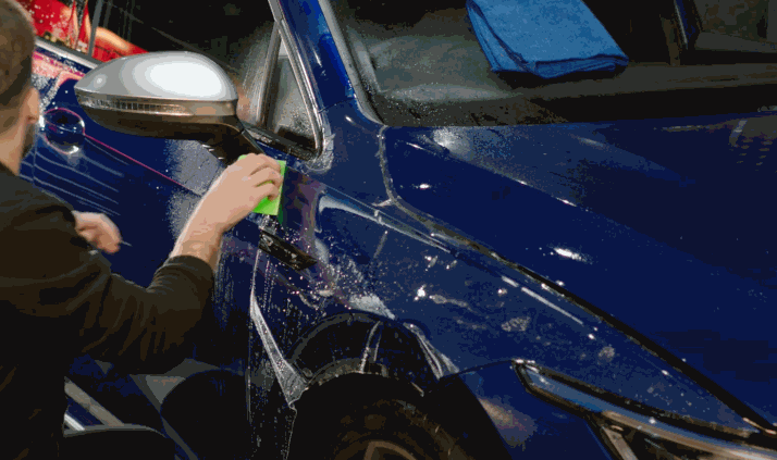 Technician applies paint protection film to the front fender of a blue car using a green squeegee while water solution spreads across the panel. The transparent paint protection film is being smoothed around the wheel arch and door seam to remove bubbles during installation.