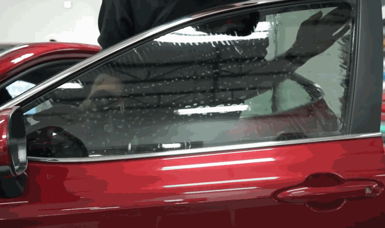 Red car door with freshly installed window tint showing water droplets during the curing process inside an auto shop.